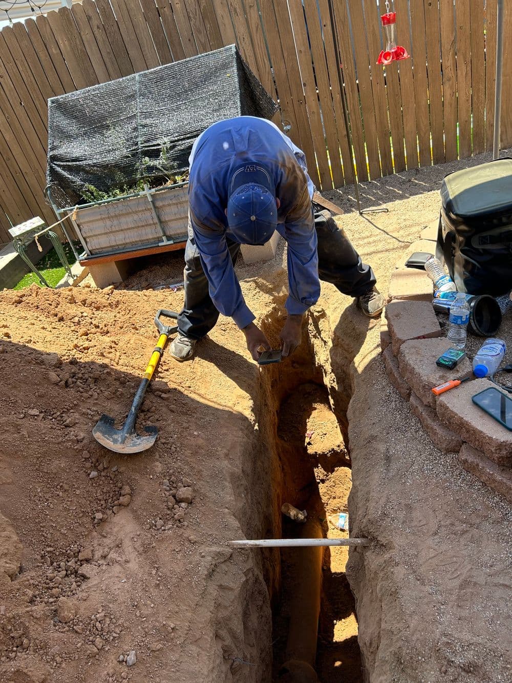 Construction worker digging a trench in a backyard, with tools and materials nearby.